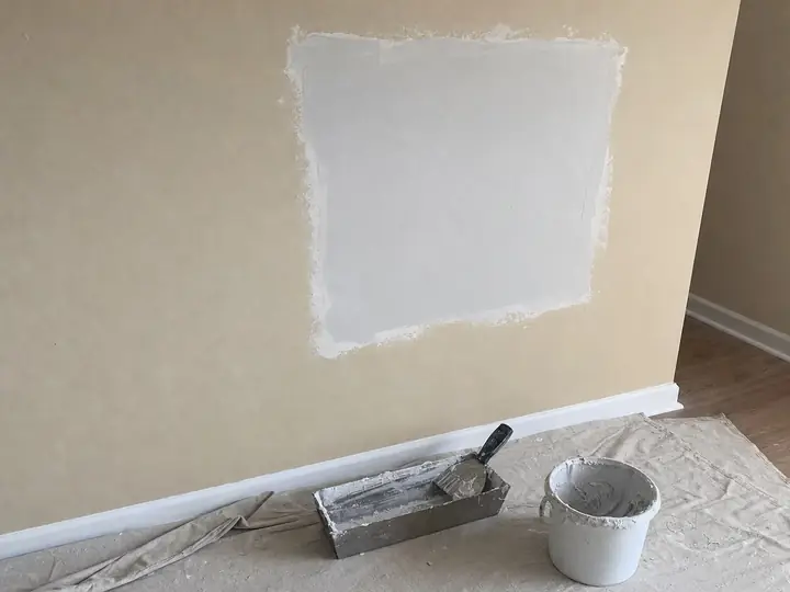 A fresh drywall patch on a beige wall, a taping knife and mud pan on a drop cloth below