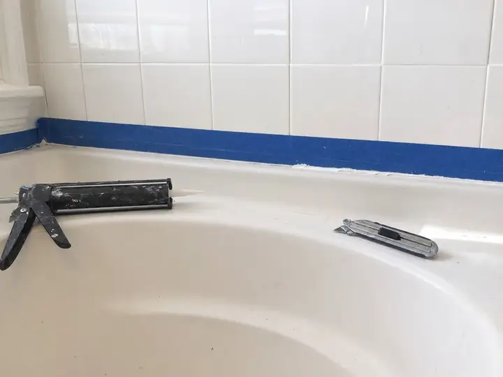 A bathtub edge taped off for new caulk, a caulk gun and a utility knife resting on the ledge