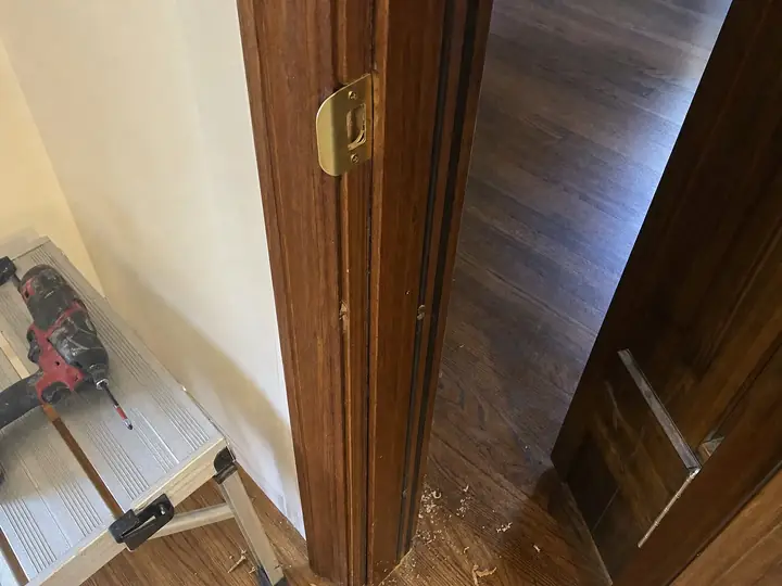 A door frame with a repaired jamb and a new brass strike plate, a cordless drill resting on a step stool